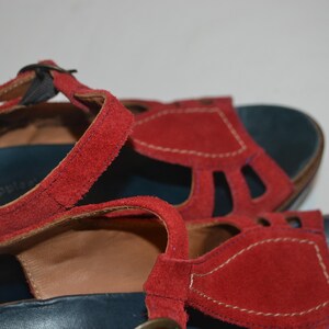 May include: Red suede sandals with a T-strap design. The sandals have a dark blue footbed and a buckle closure. The straps are stitched with white thread. The sandals are a close-up shot.