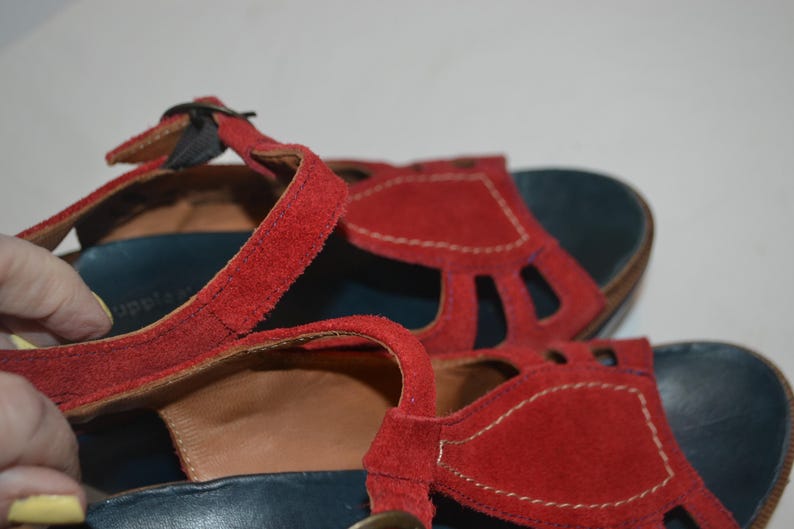 May include: Red suede sandals with a T-strap design. The sandals have a dark blue footbed and a buckle closure. The straps are stitched with white thread. The sandals are a close-up shot.