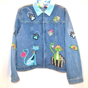 May include: Light blue denim jacket with a zip-up front and a light blue velvet collar. The jacket is decorated with embroidered cat and paw print designs, mushrooms, and umbrellas. The jacket has a whimsical, playful style.