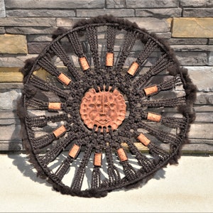 Może przedstawiać: Wiszące ozdoby ścienne z makramą w kolorze brązowym i czarnym z terakotową twarzą słońca w środku. Twarz słońca ma fakturowaną powierzchnię i jest otoczona kółkiem brązowych frędzli.