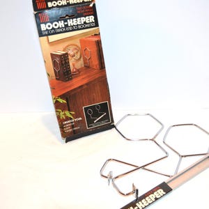 Pode incluir: Um suporte de livros de metal cromado com o texto "BOOK-KEEPER" na embalagem. O suporte de livros é projetado para manter os livros na posição vertical e apresenta um design único com múltiplas formas hexagonais.