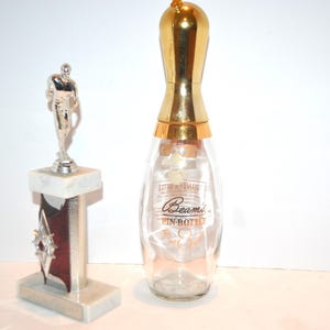 May include: A clear glass bowling pin-shaped bottle with a gold-colored top and the text "Beam's PIN-BOTTLE". A silver trophy with a football player figurine is also present.