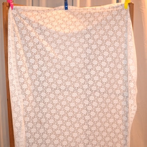 May include: A white lace fabric with a floral pattern is draped over a wooden frame and held in place with clothespins. The delicate lace has a repeating design of small flowers, creating a textured surface.