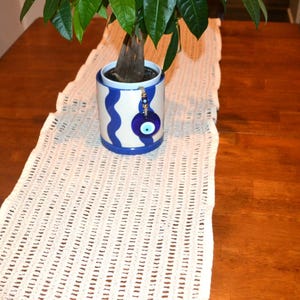 May include: A potted plant in a blue and white patterned ceramic pot with an evil eye charm, placed on a cream-colored crocheted table runner. The runner sits on a dark wooden table.