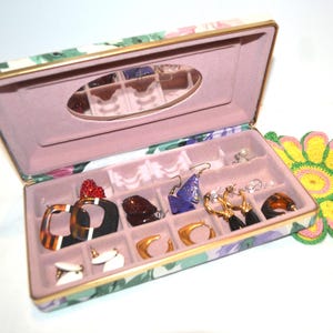 Peut inclure: Une boîte à bijoux rectangulaire avec un motif floral à l'extérieur et une doublure intérieure en velours rose. Le couvercle ouvert présente un miroir ovale, tandis que la base contient plusieurs compartiments avec diverses boucles d'oreilles.