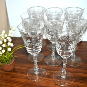 Könnte beinhalten: Eine Sammlung klarer, geätzter Weingläser. Jedes Glas hat ein Blumendesign und einen dekorativen Stiel. Die Gläser sind auf einer dunklen Holzoberfläche angeordnet, mit einer kleinen Topfpflanze im Hintergrund.