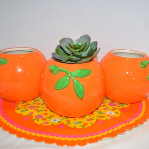 May include: Three orange ceramic planters shaped like oranges, with green leaf accents. One planter holds a succulent. The planters sit on a round, floral-patterned placemat with a scalloped edge. The overall theme is home decor.