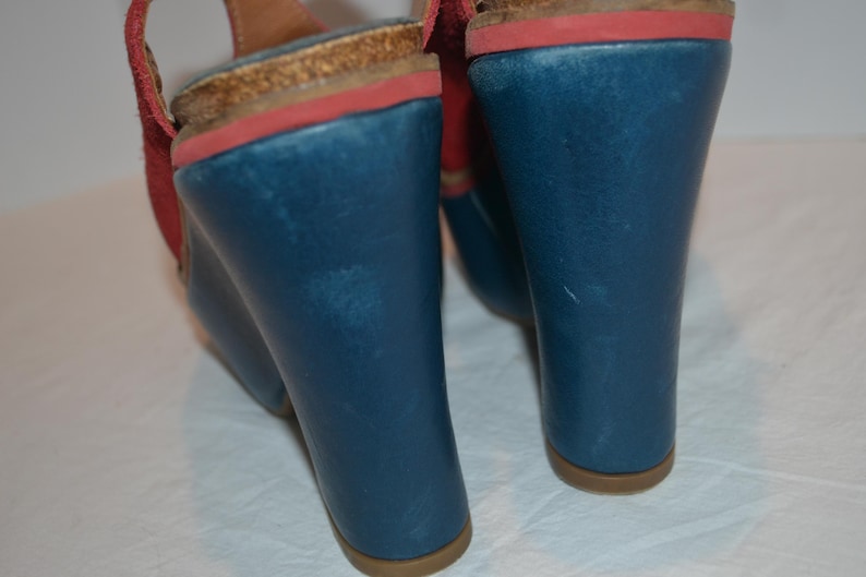 May include: Blue wedge sandals with red accents and a cork footbed. The sandals have a wedge heel and a strap. The heel height is approximately 4 inches.