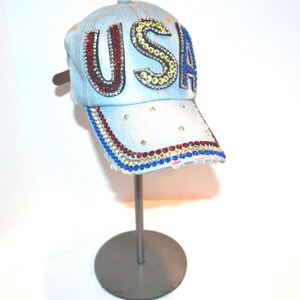 May include: Light blue denim baseball cap with the word "USA" spelled out in red, gold, and blue rhinestones. The brim is also decorated with red, gold, and blue rhinestones. The cap is displayed on a silver stand.