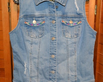 May include: A light blue denim vest with a button closure and two pockets. The vest has a rainbow-colored beaded trim around the pockets and a "d.jeans." label on the front.