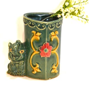 Op de afbeelding: Een groene keramische vaas met een olifantenbeeld aan de zijkant. De vaas heeft een bloemmotief met gele en rode accenten. De vaas is gevuld met witte bloemen.