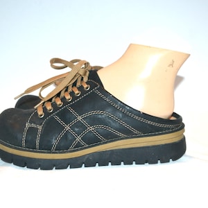 May include: A black leather mule shoe with a rounded toe and tan contrast stitching. The shoe features tan laces threaded through metal eyelets, a tan midsole stripe, and a thick, black lugged rubber sole for traction and durability.
