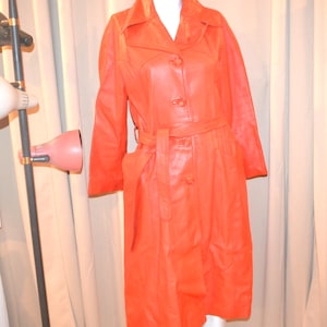 May include: A vibrant orange leather trench coat with a belted waist and button-down closure. The coat features a classic collar and long sleeves. The coat is displayed on a mannequin.
