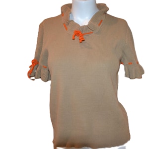 May include: A tan short-sleeved sweater with a ribbed texture. The sweater features orange accents on the ruffled collar and sleeve cuffs. The collar has a tie detail. The sweater is displayed on a mannequin.