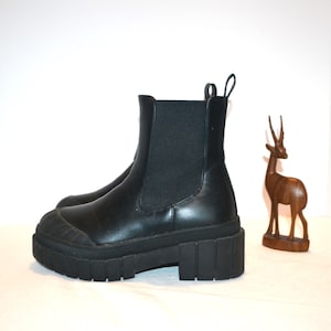 May include: Black Chelsea boots with a chunky, lug sole. The boots have a pull tab at the back and elastic side panels. A wooden deer figurine is in the background.