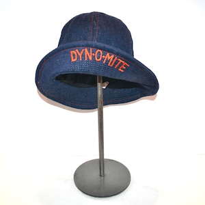 Pode incluir: Um chapéu de balde de ganga azul escuro com costuras laranja e a palavra "DYN-O-MITE" bordada na aba. O chapéu é exibido em um suporte de metal sobre um fundo branco.