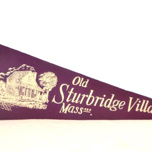 May include: A maroon pennant with a gold border and cream-colored ties. The pennant features a cream-colored illustration of a building and trees, with the words "Old Sturbridge Village Mass." in a script font.