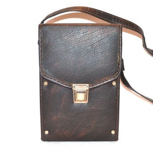 Puede incluir: Un bolso bandolera rectangular marrón oscuro con una superficie texturizada. El bolso tiene un cierre de solapa con un cierre dorado y una correa larga y delgada. Pequeños remaches dorados son visibles en la parte delantera.