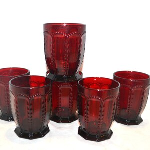 May include: Six ruby red drinking glasses. The glasses have a faceted design with a decorative pattern around the top and bottom. The glasses are arranged on a white surface.