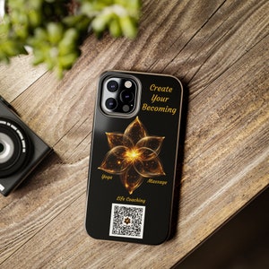 May include: Black smartphone case featuring a golden flower design with the words "Create Your Becoming". The case also includes the words "Yoga", "Massage", and "Life Coaching", along with a QR code. The case is on a wooden surface.