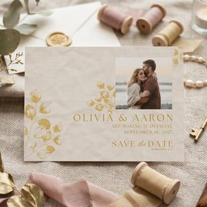 Op de afbeelding: Een save-the-date kaart met een foto van een stel, gouden bloemaccenten en de tekst "Olivia & Aaron are making it official September 18, 2027 Save the Date Temecula, CA."