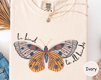May include: Ivory-colored t-shirt featuring a large butterfly design with orange, brown, and blue details. The shirt has the text "be kind to all kinds" in a handwritten style. A delicate necklace with a sun pendant is draped on the shirt.