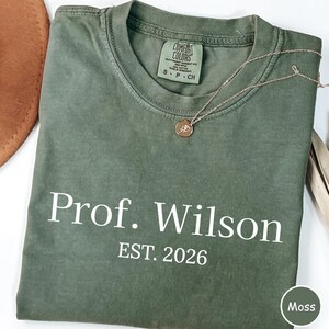 May include: A moss green t-shirt with the text "Prof. Wilson EST. 2026" in white. A delicate gold necklace with a pendant rests on the shirt. The shirt is folded and has a tag that says "Comfort Colors".