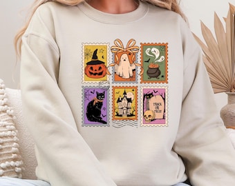 May include: Beige sweatshirt featuring six Halloween-themed postage stamp designs. The stamps depict a jack-o'-lantern, ghost, cauldron, black cat, haunted house, and a tombstone with the words "Trick or Treat".