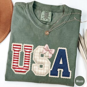 May include: A moss green t-shirt with the word "USA" appliqued on the front. The letters are in red and white stripes, gingham, and navy blue polka dots, with a pink bow and lace trim. A gold necklace is draped across the shirt.