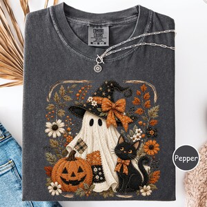 May include: A charcoal gray t-shirt featuring a graphic print of a ghost wearing a witch hat, a jack-o'-lantern, and a black cat surrounded by autumn flowers. The shirt has a Comfort Colors label at the neck and a silver chain necklace resting on it.