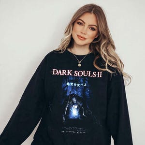 May include: A black long-sleeve sweatshirt featuring the Dark Souls II logo in white text at the top. Below the logo is a blue-toned graphic illustration of a knight in a dark forest setting with Japanese text characters printed above the figure.