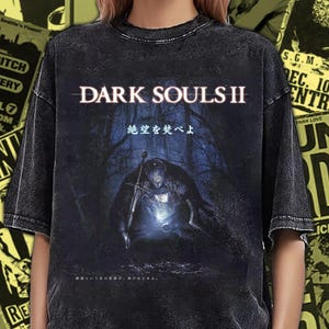 Può includere: T-shirt grigio scuro con la scritta "DARK SOULS II" e un'illustrazione di un cavaliere in una foresta oscura. La maglietta ha un aspetto vintage. Poster di concerti sullo sfondo.