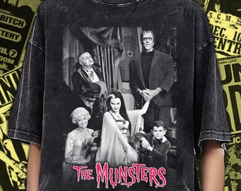 May include: A black acid-wash t-shirt featuring a black and white graphic print of the television show cast The Munsters. The print includes the characters Herman, Lily, Grandpa, Marilyn, and Eddie, with the title The Munsters in pink text below.