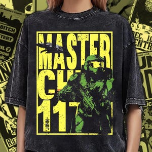 Pode incluir: Uma t-shirt preta com efeito de lavagem ácida, apresentando um grande gráfico retangular amarelo. A estampa exibe o texto MASTER CHIEF 117 e uma ilustração de um soldado com armadura verde segurando uma arma, com o nome JOHN impresso na vertical.