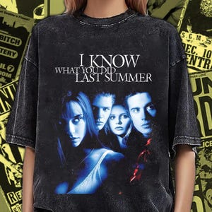 Könnte beinhalten: Schwarzes, übergroßes T-Shirt mit dem Filmtitel "I Know What You Did Last Summer" und einem blau getönten Bild von Schauspielern. Das Shirt hat einen Vintage-Look. Konzertplakate bilden den Hintergrund.