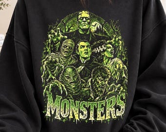 Puede incluir: Una sudadera negra de cuello redondo con un estampado gráfico en verde vibrante de personajes clásicos de películas de terror, incluyendo a Frankenstein, una momia, un vampiro, una novia, una criatura del pantano y un hombre lobo, con la palabra MONSTERS.