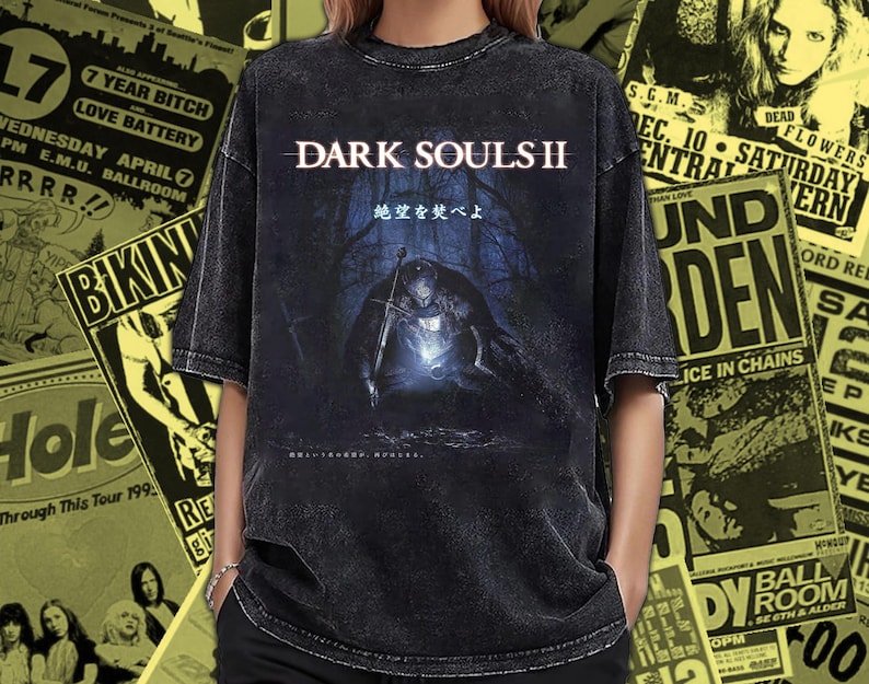 May include: Dark gray t-shirt featuring the text "DARK SOULS II" above an illustration of a figure with a sword in a dark forest. Japanese text is also present. The shirt has a vintage, distressed look.