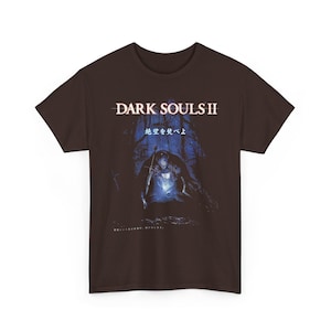 May include: Brown t-shirt featuring the text "DARK SOULS II" above an illustration of a knight in a dark forest. The graphic is in shades of blue and white, with Japanese text below the main title.