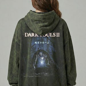 May include: Green hooded sweatshirt with a faded, distressed look. The back features the text "DARK SOULS II" above a graphic of a knight in a dark forest. Additional Japanese text is also visible.