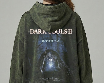 Puede incluir: Sudadera con capucha verde con un aspecto descolorido y desgastado. La parte trasera presenta el texto "DARK SOULS II" sobre un gráfico de un caballero en un bosque oscuro. También se ve texto japonés.