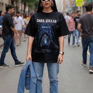 May include: Black graphic t-shirt with the text "DARK SOULS II" and a dark forest scene. The person is wearing light blue jeans, black shoes, white sunglasses, and holding a blue denim jacket.