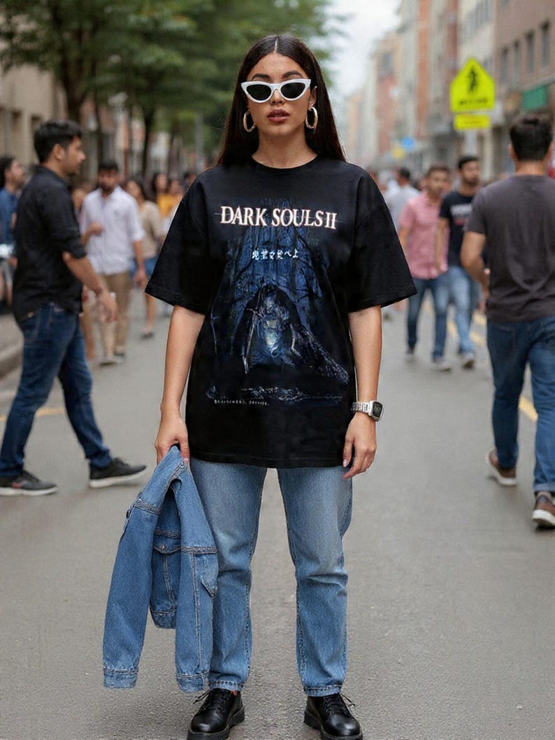 May include: Black graphic t-shirt with the text "DARK SOULS II" and a dark forest scene. The person is wearing light blue jeans, black shoes, white sunglasses, and holding a blue denim jacket.