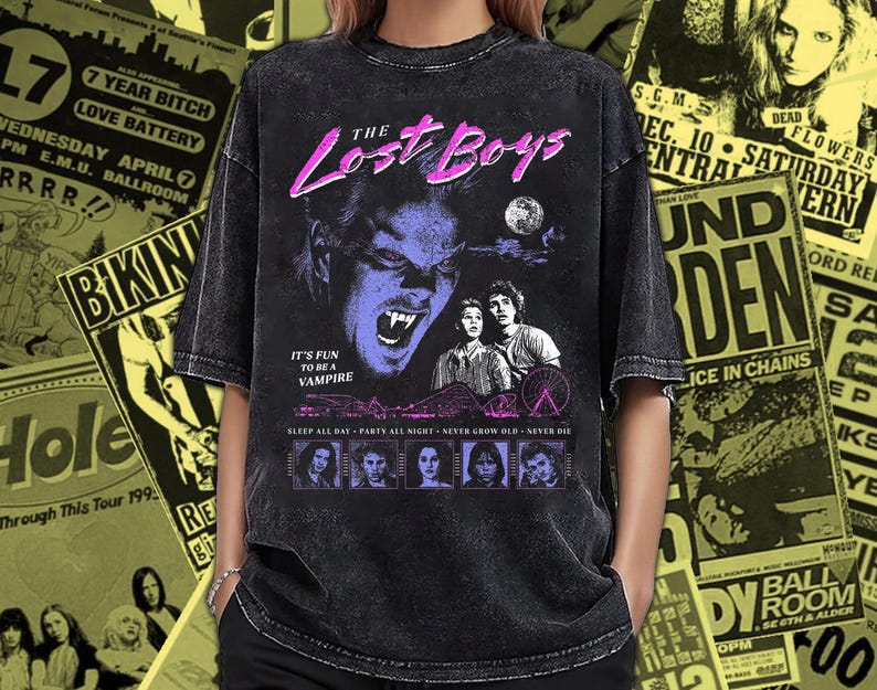 May include: A black acid-wash t-shirt featuring a graphic print from the movie The Lost Boys. The design includes a vampire face, the text The Lost Boys, It's fun to be a vampire, Sleep all day, Party all night, Never grow old, Never die, and characters.