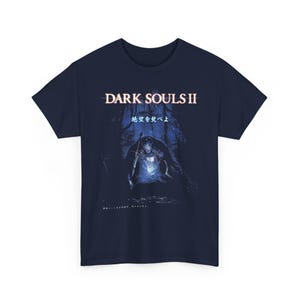 May include: Navy blue t-shirt featuring the text "DARK SOULS II" above an illustration of a knight holding a sword in a dark forest. Japanese text is also present. The design is in shades of blue and white.