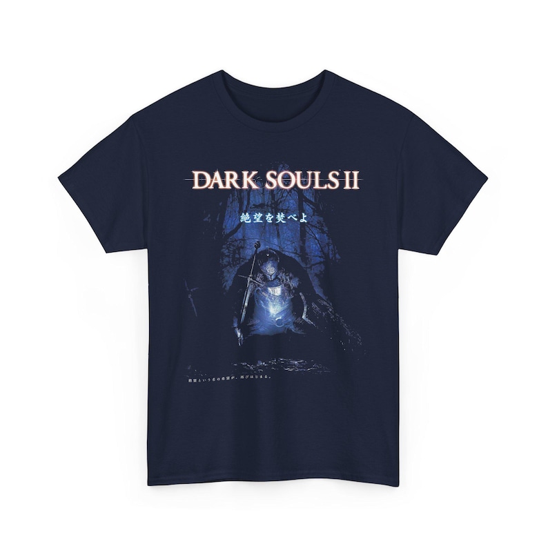 May include: Navy blue t-shirt featuring the text "DARK SOULS II" above an illustration of a knight holding a sword in a dark forest. Japanese text is also present. The design is in shades of blue and white.
