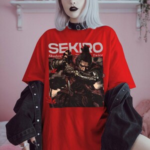 Può includere: T-shirt rosso con il titolo del gioco "SEKIRO" in bianco. La maglietta presenta una grafica di un guerriero in azione. La persona indossa una giacca di jeans nera sopra.