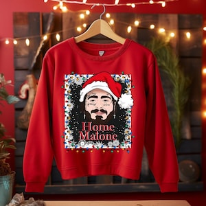 May include: A red sweatshirt featuring a graphic of a man wearing a Santa hat, surrounded by Christmas lights. The text "Home Malone" is below the graphic. The sweatshirt is hanging on a wooden hanger.