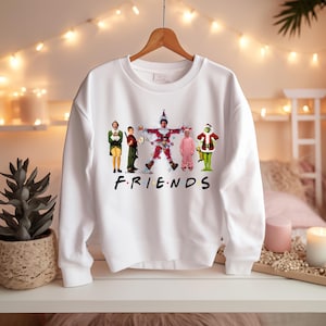 Puede incluir: Sudadera blanca de cuello redondo con un gráfico de personajes de las películas "Elf" y "The Grinch" al estilo de la serie de televisión "Friends". La palabra "FRIENDS" está impresa en letras negras en la parte inferior del gráfico.