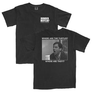 May include: Black t-shirt featuring the text "Dunder Mifflin Scranton Hardcore" on the front. The back of the shirt displays a black and white image of a man with the text "WHERE ARE THE TURTLES? WHERE ARE THEY??"