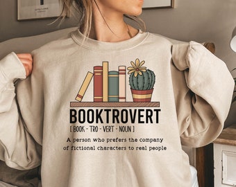May include: Beige sweatshirt with a graphic design of a bookshelf with books and a cactus. The text "BOOKTROVERT" is written in black with the definition "[BOOK-TRO-VERT - NOUN] A person who prefers the company of fictional characters to real people."